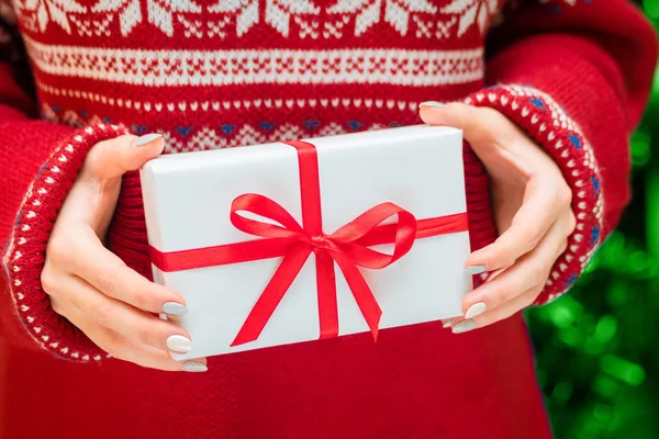 Nő mutatja gyönyörű fehér ajándék doboz piros szalag-Vértes jelen holiday pulóver háttér és a karácsonyfa. — Stock Fotó