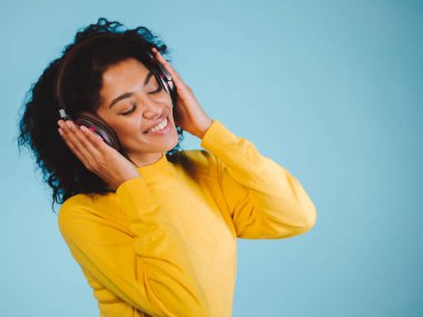 Sarı kazak mavi bir arka plan üzerinde duran Kapalı gözlerle gülümseyen güzel genç kadın, müzik kulaklık