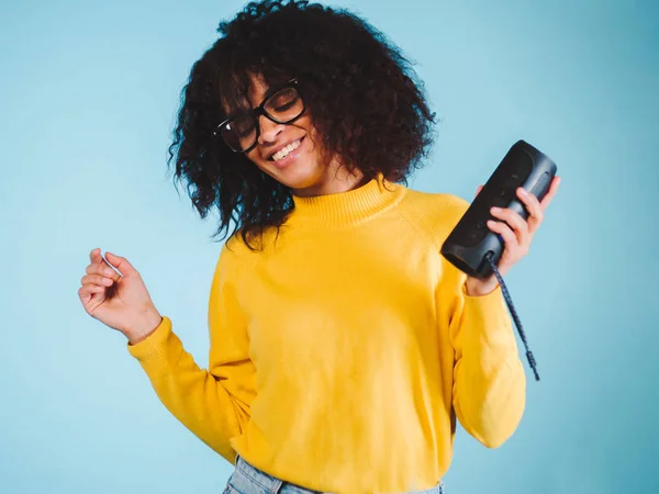 Junge schöne afrikanische Frau genießt und tanzt vor blauem Hintergrund. moderne trendige Mädchen mit Afro-Frisur hören Musik von Bluetooth tragbaren Lautsprecher. — Stockfoto