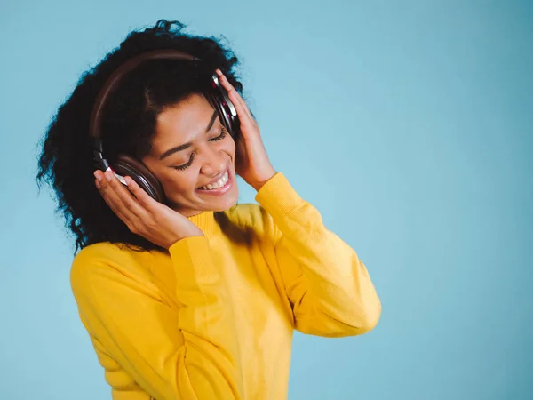 Красива молода жінка в навушниках слухає музику, посміхається закритими очима, стоячи на синьому фоні в жовтому светрі — стокове фото