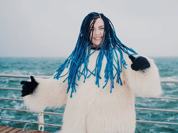 Stylisches Hipstermädchen in weißem Pelzmantel und blauen Kanekalon-Zöpfen, das sich im Winter am Strand in der Nähe des Meeres amüsiert. gefärbte Haare, trendige Frau. — Stockfoto