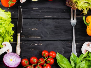 Taze çiftlik sebze, sarı kırmızı biber biber, fesleğen, domates, mantar, kırmızı soğan, limon, marul ile mutfak siyah çerçeve arka plan. Gıda üstten görünüm. Bıçak ve çatal orta