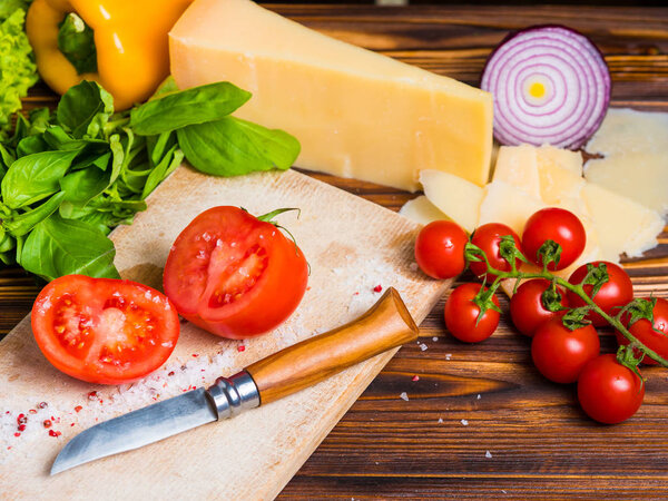 Appetizing still life. Fresh bulgarian yellow pepper, cherry tomatoes, cheese, lettuce leaves, red onion, green basil on wooden board. The knife for cutting vegetables. Top view