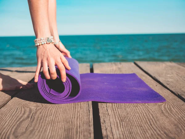 Giovane bella donna sportiva che si prepara per lo yoga asana sulla spiaggia di mare vicino all'acqua. Ragazza che copre stuoia viola. Concetto sanitario . — Foto Stock