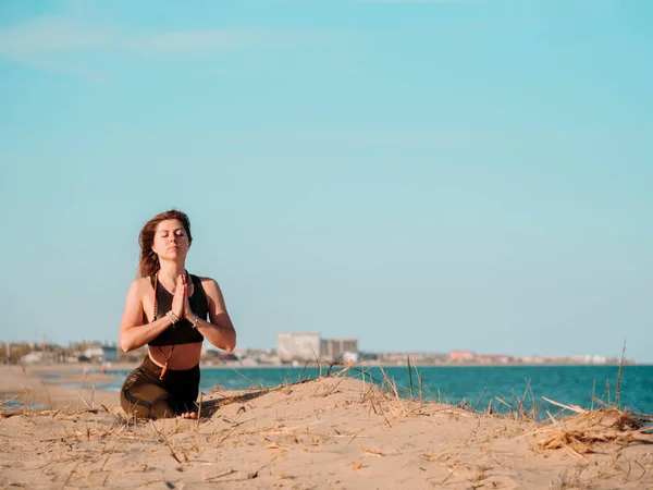 Young beautiful sporty woman doing yoga asana on sea beach near water. Girl practicing exercises. City background. Health concept. Copy space. — Stock Photo, Image