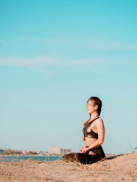 若い美しいスポーティな女性の水の近くの海のビーチでヨガのアーサナを行います。女の子の瞑想。背景の都市。健康の概念。コピー スペース. — ストック写真