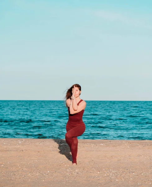 Молода красива спортивна жінка в червоному костюмі робить йогу асану на морському пляжі біля води. Дівчина практикує вправи. Концепція здоров'я . — стокове фото