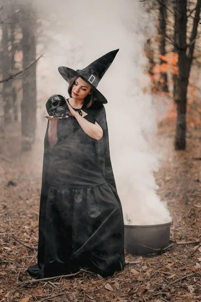 Bruja negra sosteniendo el cráneo en las manos, acariciándolo sobre el fondo del bosque de otoño. Horror, halloween, vacaciones cosplay, concepto mágico — Foto de Stock