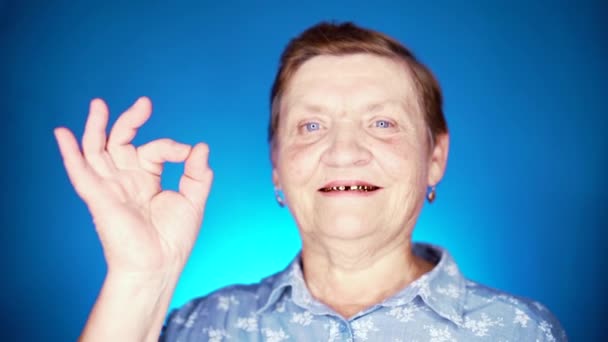 Piękny portret uśmiechniętej starszej kobiety na niebieskim tle. Biała babcia patrząc w aparat i pokazując znak ok - gest aprobaty. — Wideo stockowe