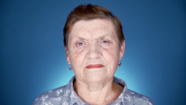 Una abuela caucásica mirando a la cámara. Retrato de mujer anciana sonriente en estudio sobre fondo azul. — Vídeos de Stock