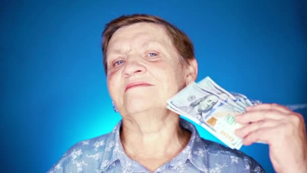 Babcia się uśmiecha, cieszy się, że dostaje zasiłki papierową walutą. Emerytka w podeszłym wieku na niebieskim tle z dolarami. — Wideo stockowe