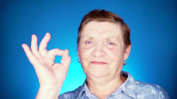 Чудовий портрет усміхненої літньої жінки на синьому тлі. Кавказька бабуся дивиться на фотоапарат і показує чіткий жест схвалення.. — стокове відео