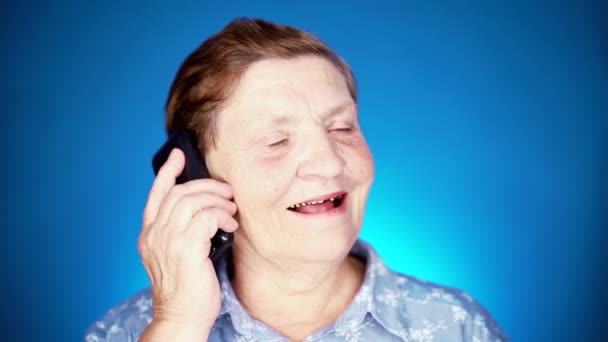 Пожилая женщина разговаривает с мобильным устройством на голубой стене студии. Бабушка улыбается, приятно беседует. замедленное движение — стоковое видео