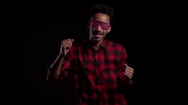 Hombre africano con gafas de neón brillantes moviéndose al ritmo de la música en el fondo del estudio negro. Bailando tipo cómico positivo. Movimiento lento . — Vídeos de Stock