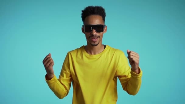 Hombre africano con gafas de neón brillantes moviéndose al ritmo de la música en el fondo del estudio. Bailando tipo cómico positivo. Movimiento lento . — Vídeos de Stock