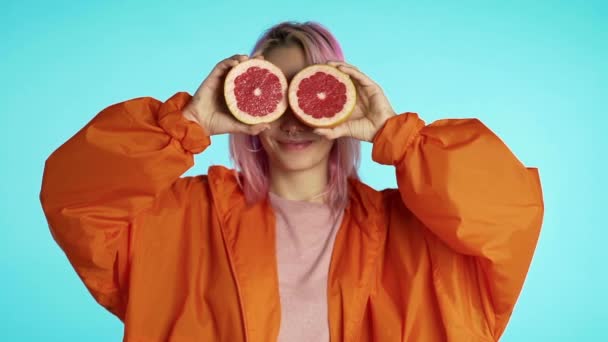 Radostný mladý hipster s obarveným růžovým účesem se dvěma půlkami šťavnatého grapefruitu na očích. Zdravý životní styl a pojetí stravování. Zpomalit záběry z modrého studia. — Stock video