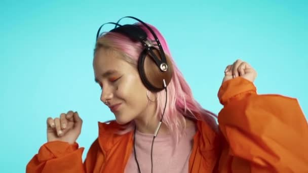 Femme hipster avec une apparence extraordinaire dans les écouteurs en imperméable orange sur fond bleu. Jolie fille aux cheveux roses aime écouter de la musique . — Video