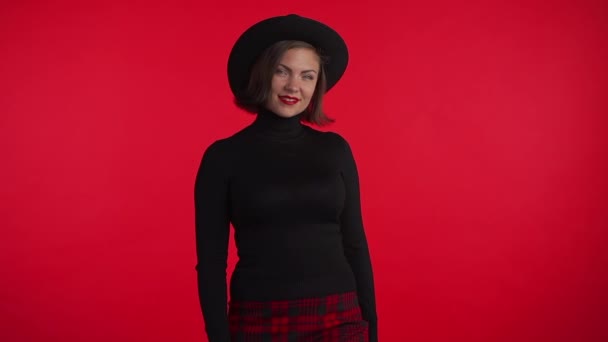 Portrét mladé hezké ženy na červeném pozadí ve studiu. Trendy hipster dívka v černém klobouku a kostkované mini sukně. Usmívající se dáma. — Stock video