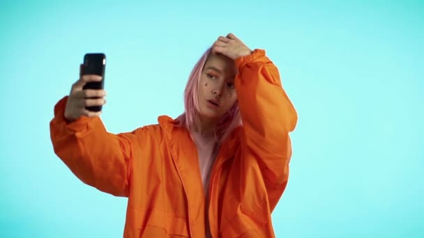 Dziewczyna z różowymi włosami i niezwykłym wyglądem zrobić selfie na niebieskim tle. Korzystanie z nowoczesnych technologii - smartfon, portale społecznościowe — Wideo stockowe