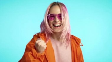 Mavi arka plandaki heyecanlı, mutlu hipster 'a şaşırdım. Pembe saçlı ve olağanüstü görünüşlü bir kız evet zafer jesti gösteriyor, başarıya ulaştı, hedeflere ulaştı.. 