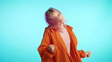 Pembe saçlı, eğlenceli, gülümseyen, stüdyoda mavi arka planda dans eden tuhaf bir kadın. Müzik, dans konsepti, ağır çekim