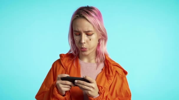 Девушка с необычным внешним видом играет в игру на смартфоне на голубой стене студии. Использование современных технологий - приложения, социальные сети — стоковое видео