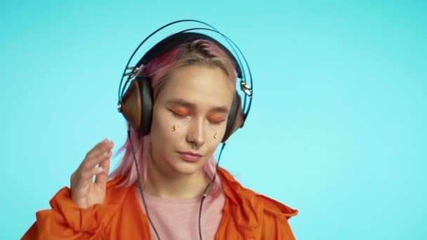 Mooi meisje met roze haar luistert graag naar muziek. Hipster vrouw met buitengewone verschijning in oranje regenjas op blauwe achtergrond. — Stockvideo