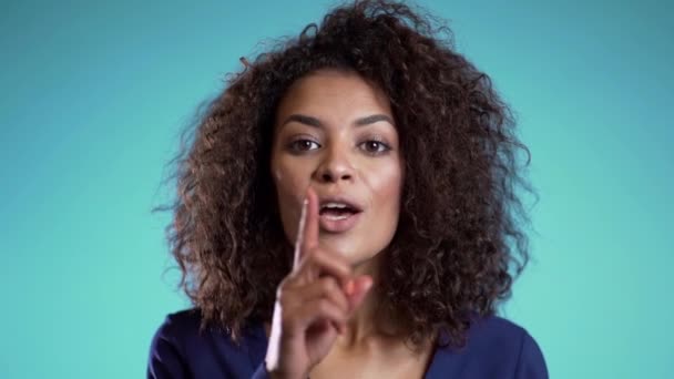 Mujer afroamericana sosteniendo un dedo en sus labios sobre fondo azul. Gesto de silencio, secreto, silencio. De cerca. . — Vídeo de stock