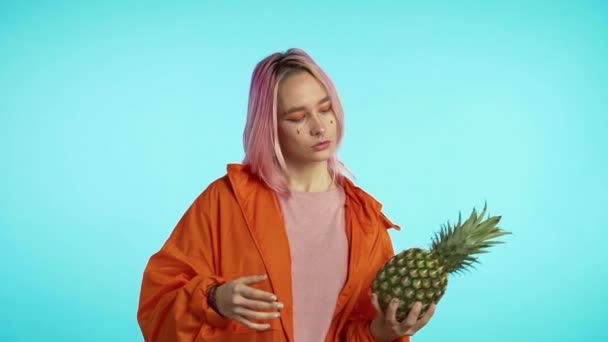 Playful young hipster with dyed pink hairstyle throws hand to hand fresh pineapple. Healthy exotic lifestyle and eating concept. Slow motion blue studio footage. — Stock Video