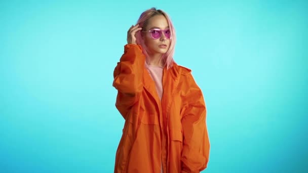 Retrato de linda chica peculiar en estudio azul. Imágenes coloridas vibrantes. Mujer Hipster en abrigo naranja con peinado rosa. Movimiento lento . — Vídeos de Stock