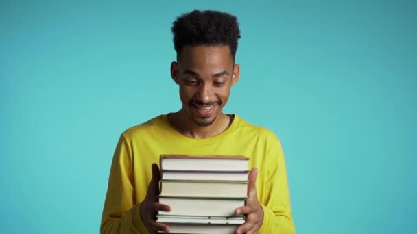 Afrikansk student på blå bakgrund i studion håller stack med universitetsböcker från biblioteket. Killen ler, han är glad att ta examen. — Stockvideo