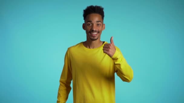 Hezký černoch ve žlutém oblečení na modrém pozadí studia se usmívá na kameru a dává palce nahoru. Šťastný chlapík projevující souhlas. Vítěz. Úspěch. — Stock video