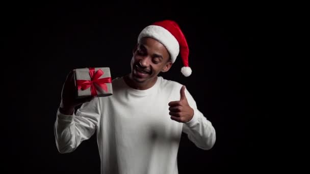 サンタの帽子のハンサムな男はクリスマスプレゼントを持っていて、黒いスタジオの背景で踊ることはカメラに笑顔を見せ、親指を上げます。幸せなアフリカ系アメリカ人だスローモーション. — ストック動画