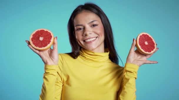青いスタジオの背景にジューシーなグレープフルーツの2つの半分と黄色のきれいな女性。健康的な食事、食事、抗酸化物質の概念. — ストック動画