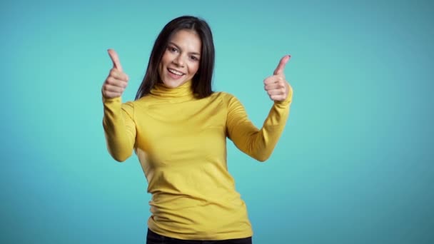 Vencedor. Sucesso. Menina morena em amarelo desgaste no fundo azul sorri para a câmera e dá polegares para cima. Mulher feliz mostrando gesto positivo . — Vídeo de Stock