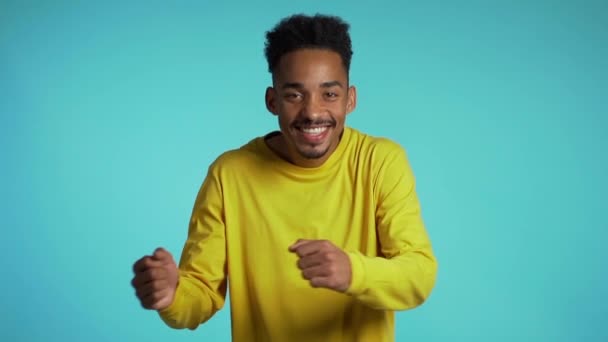 Olumlu gülümseyen adam mutluluğu, uyumu ve eğlenceyi resmediyor. Sarı elbiseli Afrikalı adam mavi stüdyo arka planında dans ediyor. Yavaş çekim. — Stok video