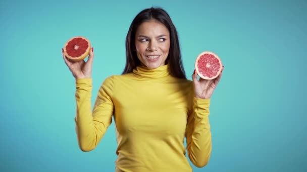 Aranyos lány pózol két fél szaftos grapefruittal, kék stúdió háttérrel. Egészséges táplálkozás, diéta, antioxidáns fogalom. Lassú mozgás.. — Stock videók