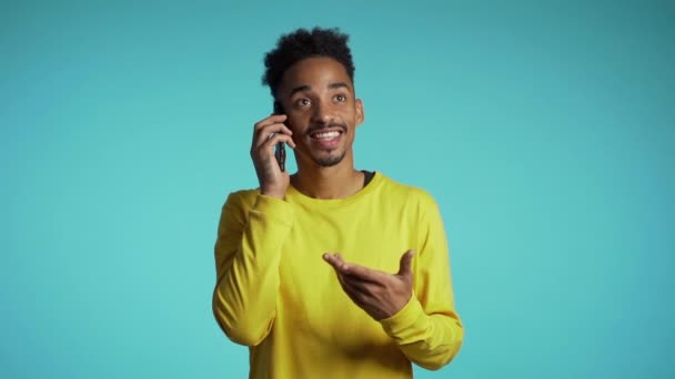 Homem americano africano sorridente em amarelo, a falar no telemóvel sobre fundo azul. Bonito homem segurando e usando telefone inteligente. Cara com tecnologia — Vídeo de Stock