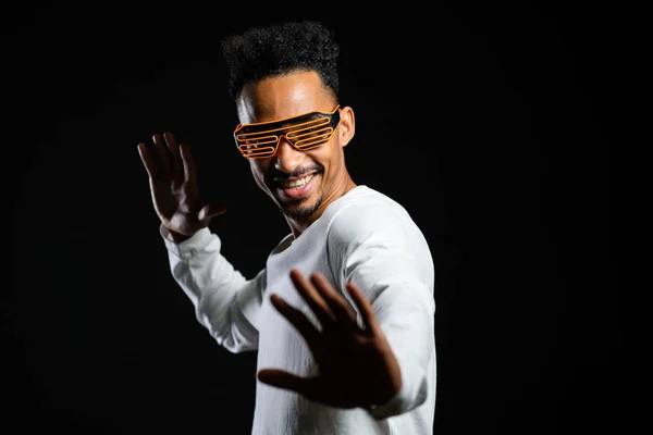Hombre africano con gafas de neón brillantes moviéndose al ritmo de la música en el fondo del estudio negro. Bailando chico cómico positivo . — Foto de Stock