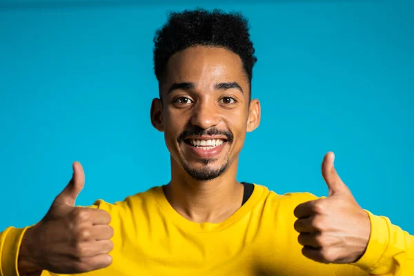 Bonito homem preto em amarelo desgaste no fundo do estúdio azul sorri para a câmera e dá polegares para cima. Um tipo feliz a mostrar um gesto de aprovação. Vencedor. Sucesso . — Fotografia de Stock