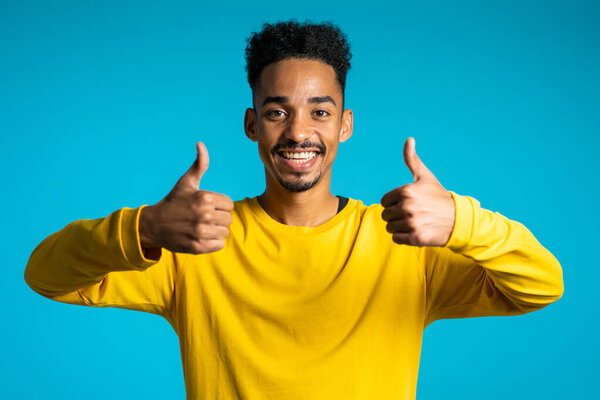 Handsome black man in yellow wear on blue studio background smiles to camera and gives thumbs up. Happy guy showing gesture of approval. Winner.Success.