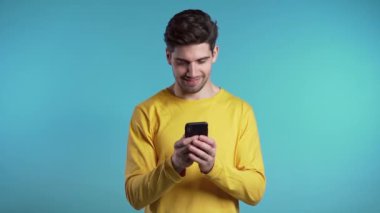 Genç adam gülümsüyor ve mavi arka planda cep telefonu kullanıyor. Yakışıklı Avrupalı adam elinde tutuyor ve akıllı telefonla mesaj atıyor. Teknolojili çocuk..
