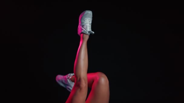 Mladá žena tanečnice na černém pozadí s neonovým světlem pohybující její sexy nohy v bílých teniskách. Úžasný tanec. Na konci roztáhne nohy do provazu. — Stock video