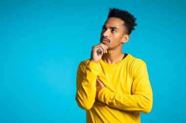 Düşünen adam mavi arka planda etrafa bakıyor. Dalgın yüz ifadeleri için endişelenmiş. Kıvırcık saçlı Afrikalı Amerikalı erkek model kararlar alıyor, fikir sayıyor.