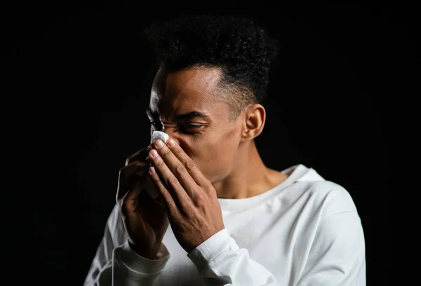Un joven afroamericano estornuda en el tejido. El hombre aislado está enfermo, resfriado o tiene una reacción alérgica. Salud, medicina, enfermedad, concepto de tratamiento — Foto de Stock
