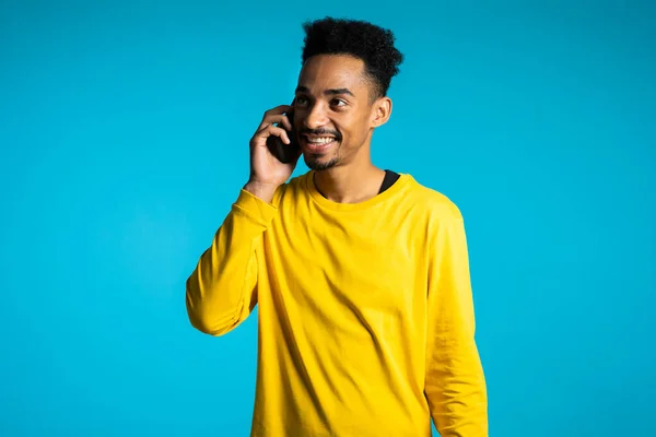 Hombre afroamericano sonriente vestido de amarillo hablando por teléfono móvil sobre fondo azul. Hombre guapo sosteniendo y usando un teléfono inteligente. Chico con tecnología — Foto de Stock
