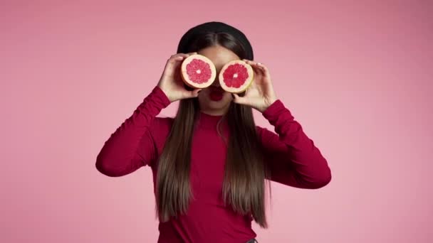 在粉红的画室背景上画着一位身穿红衣服的年轻漂亮女子的画像，她有一半的柚子汁。 健康饮食、节食、抗氧化剂概念 — 图库视频影像