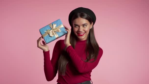 ピンクの壁の背景に弓と青のギフトボックスを保持完璧なメイクを持つ美しい若い女性。女の子は笑って、彼女はプレゼントを得るために満足している. — ストック動画