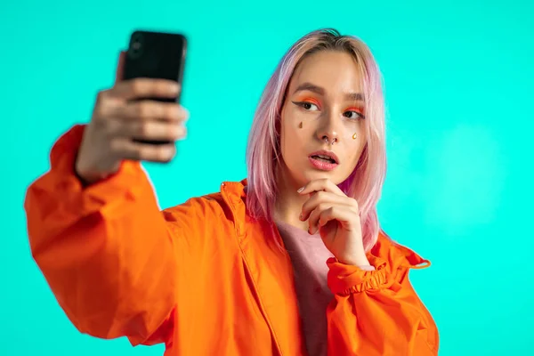 Menina com cabelo rosa e aparência extraordinária fazer selfie no fundo azul. Usando a tecnologia moderna smartphone, redes sociais — Fotografia de Stock