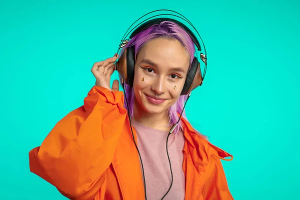 Το όμορφο κορίτσι με τα ροζ μαλλιά απολαμβάνει να ακούει μουσική. Hipster γυναίκα με εξαιρετική εμφάνιση σε πορτοκαλί αδιάβροχο σε μπλε φόντο. — Φωτογραφία Αρχείου
